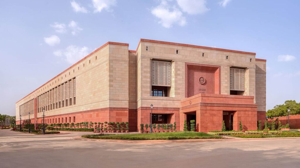 Modern red sandstone facade of India's New Parliament House in New Delhi under clear blue skies, showcasing grand central entrance and expansive architecture designed for 888 MPs – key context for the failed 131st Amendment seat expansion debate.